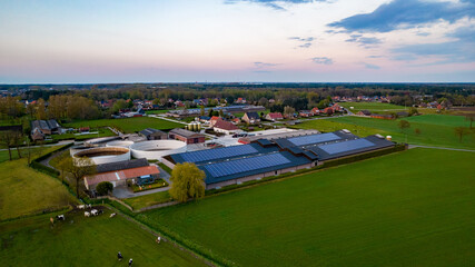This aerial shot captures a contemporary farming complex at dusk, as the days last light paints the scene with soft hues. The farm features a range of buildings with modern designs, including circular