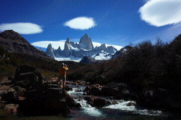 Hiking Patagonia Mount Fitz Roy, a man photographs towering granite peaks and unusual cloud formations while standing on rocks next to a river in Los Glaciares National Park in southern Argentina