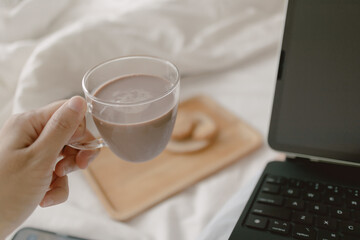 Image of woman hand holding and drinking coffee, milk tea cup while working and using laptop on white bed, freelancer lifestyle.