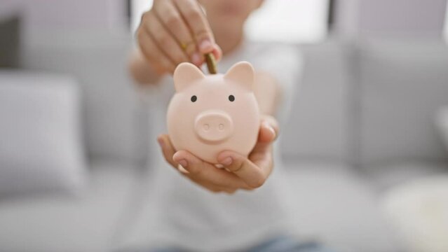 Adorable blond boy sitting comfortably on sofa, confidently inserting coin in piggy bank while enjoying relaxed lifestyle at home. boy's joyful expression reveals positive finance learning.