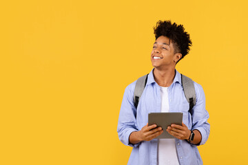 Optimistic black male student with tablet looking up at free space on yellow