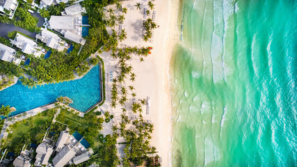 Aerial view of a tropical beach resort with a large swimming pool, palm trees, and turquoise ocean, conveying a concept of luxury travel or vacation