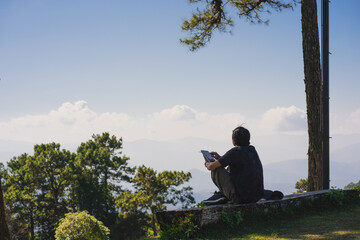 Fototapeta premium landscape and travel concept with solo freelancer man sit on wooden and use tablet work from outdoor with layer of mountain background