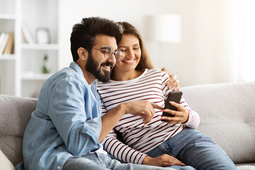Happy indian couple sitting on sofa at home, using smartphone