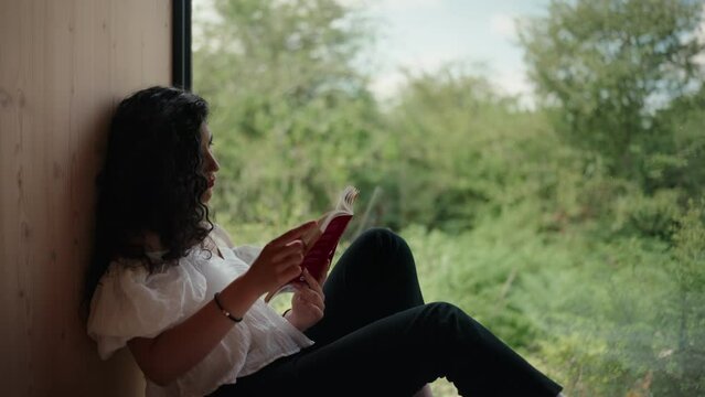 Woman reading a book by window in a cabin in nature medium shot 02