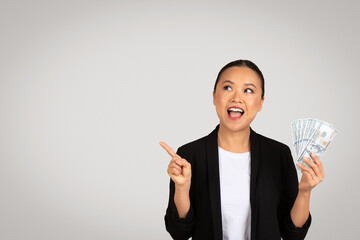 Happy surprised young asian woman in suit hold money, point finger at empty space