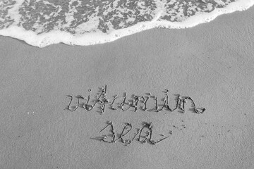 Words Vitamin Sea written on wet beach sand beside foamy wave, black and white
