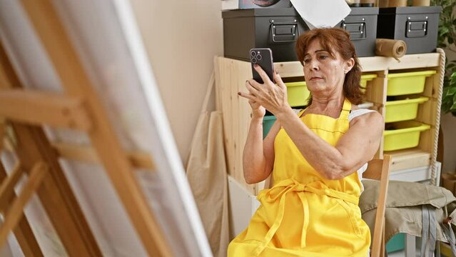 Mature artist woman in yellow apron takes photo with smartphone, focusing on her art in a creative studio.
