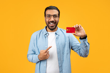 A cheerful man in a denim shirt holding a red credit card and pointing at it with his other hand