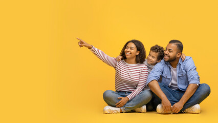 Happy African American Family Of Three Sitting On Floor And Pointing Aside