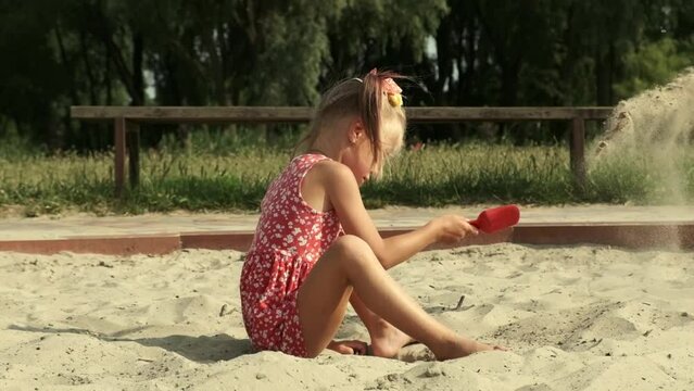 Cute little girl plays in the sandbox with a shovel wearing red dress during hot sunny summer day