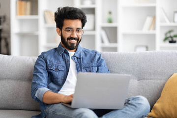 Indian man wearing glasses using laptop while sitting on couch at home