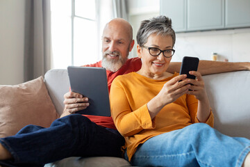 Happy Senior Spouses Relaxing With Modern Gadgets On Couch At Home