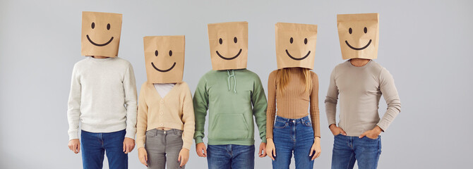 Portrait five casual male and female people with paper shopping package bags over heads with happy smiling emoticon mouth eyes facial expression drawn on them standing on gray color studio background - obrazy, fototapety, plakaty