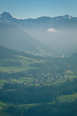 Fototapeta na wymiar Alpenlandschaft, Baum und wälder. Dorf mit Kirche, nebel und Berggipfel. Schnee auf den Bergen. Felder und wiesen. Blick ins Tal