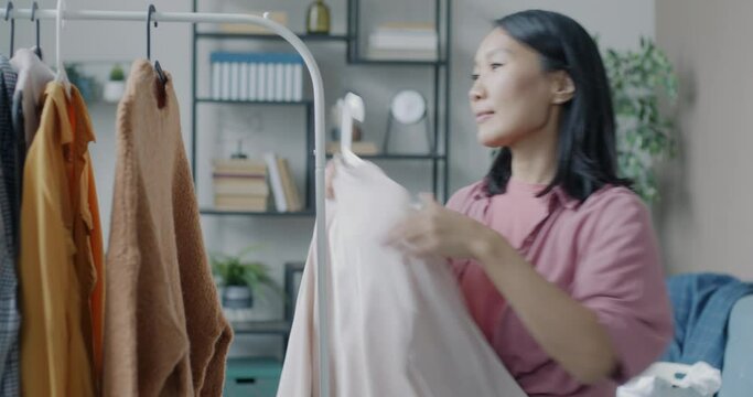 Attractive Asian girl hanging clothes on rails hanger smiling doing housework looking at garments collection. Modern lifestyle and fashion concept.