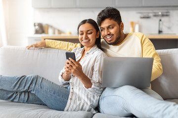 Engaged indian couple relaxing on couch with smartphone and laptop