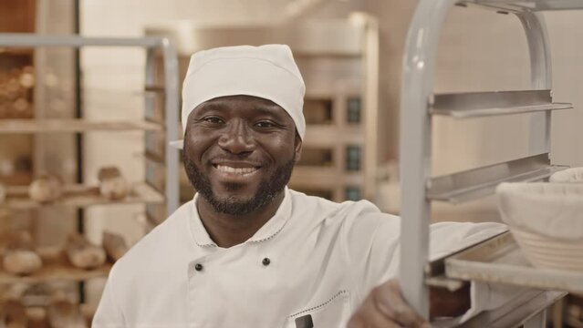 Chest up slowmo portrait of young African American baker wearing white chef uniform smiling at camera while leaning on tray rack with bread baskets in bakery