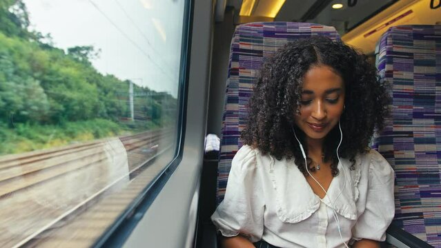 Front view of young businesswoman commuting sitting by window on moving train working on laptop and listening to music or podcast on earphones - shot in slow motion