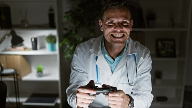 Smiling hispanic male doctor in lab coat enjoys gaming with a controller in a dark clinic office.
