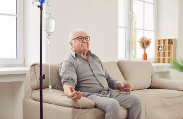 Senior man sitting on the couch at home or in clinic while receiving IV drip infusion and vitamin therapy in his blood. Smiling elderly male person in casual clothes receiving injection therapy. - obrazy, fototapety, plakaty