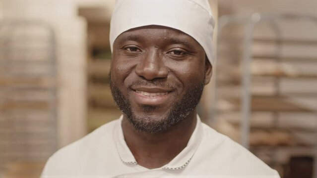 Medium closeup with slowmo portrait of young Black male chef in workwear looking at camera standing in bakery kitchen with empty tray racks in blurred background