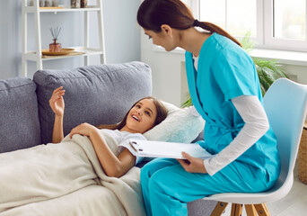Doctor visiting sick child at home. Young woman pediatrician in blue green scrubs holding clipboard, sitting on chair by couch and talking with little girl patient who is lying under blanket