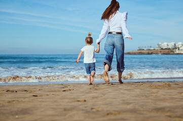 Rear portrait of a Caucasian young mother and her little child girl holding hands while walking...
