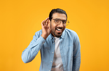 A delighted man in a blue denim shirt with glasses cupping hand near ear in a listening gesture