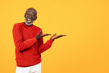 Cheerful elderly Black man in red sweater showcasing with hands on an empty space