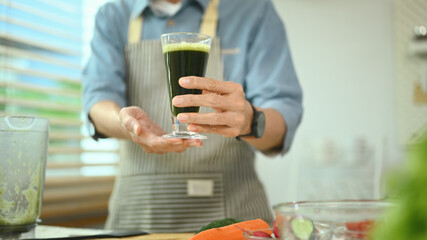 Healthy senior man holding a glass of green vegetable smoothie near ingredients in kitchen