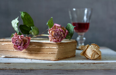 Dried roses lie on an old book. In the background is a glass of red wine.