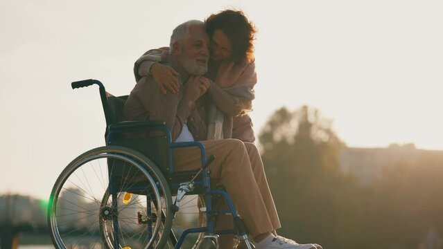 Wife walking with husband in wheelchair. Senior family couple spending time in city park near pond. Woman hugging man showing love care, they talking smiling. Living with physical disability concept.