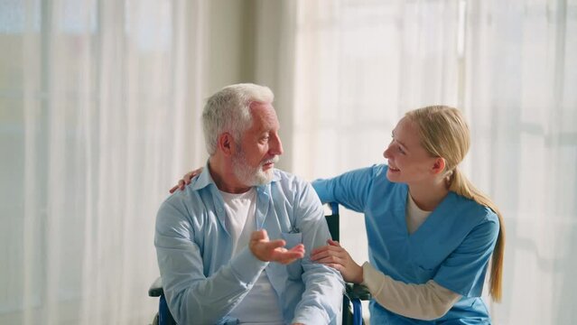 Nurse talking senior man in wheelchair. Female social welfare physical therapist looks after senior male, listening, providing medical care at home. Living with legs paralysis, special needs people.