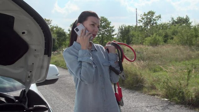 Frustrated, the driver angrily communicates with the assistance service over the cell phone, holds jumpstart ignition cables in hand. car troubles with the service provider.