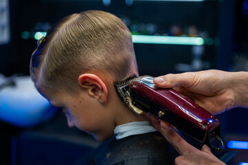 Cute emotional fair-haired smiling boy with blue eyes at the barber shop. Stylist's hands with tools. Children's fashion. Indoors, dark background, neon. Selective focus
