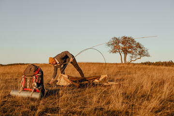 Male backpacker setting up camping tent at campsite in field at sunset in autumn 