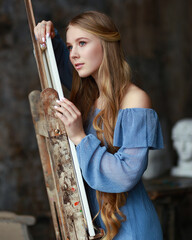 Beautiful blonde woman artist with long hair in blue vintage dress leaning on canvas. Romantic...