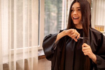 Smiling beautiful girl holds scissors and bun of her cut hair in her hands - obrazy, fototapety, plakaty