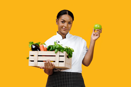 Cheerful black female chef holding green apple and crate of vegetables