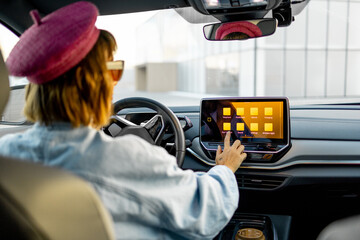 Woman use a touchscreen while driving modern electric car at city. Stylish woman traveling by car
