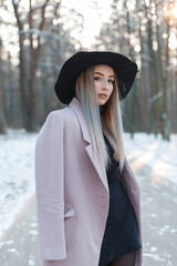 Elegant beautiful natural girl in fashionable clothes with a black hat and dress with a purple coat posing in the park on a winter snowy day