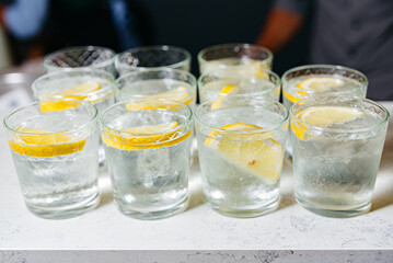 Chilled Lemon Water Glasses on Bar Counter. Multiple glasses of refreshing ice-cold lemon water garnished with lemon slices, ready to be served on a bar counter.