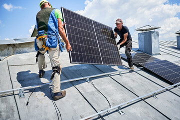 Workers building solar panel system on metal rooftop of house. Two men installers carrying...