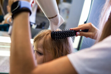 Hair salon, close-up of hairdresser woman makes combing hair dryer for cute little girl, haircare....