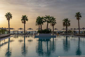 Fototapeta na wymiar Sunrise over the pool and sunbeds. On the background of the sea coast. Big palm trees.