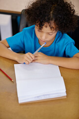 Student, thinking and child writing in notebook above desk with pencil, paper and learning in...
