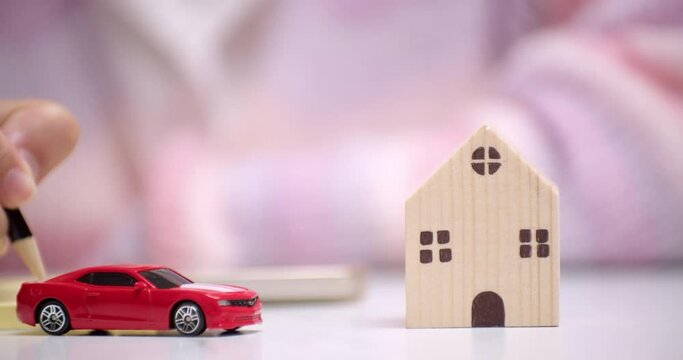 Close-up of a hand holding a pencil and writing something on a yellow paper, behind a red toy car that signifies car loan financing.