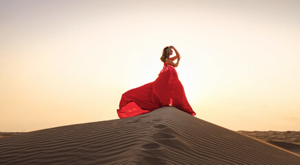 Desert adventure. Young arabian Woman in red silk dress in sands dunes of UAE desert at sunset,...