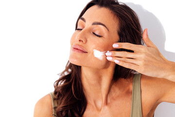 Beautiful caucasian woman smears her cheeks with cream. Young brunette female smiles with her eyes closed